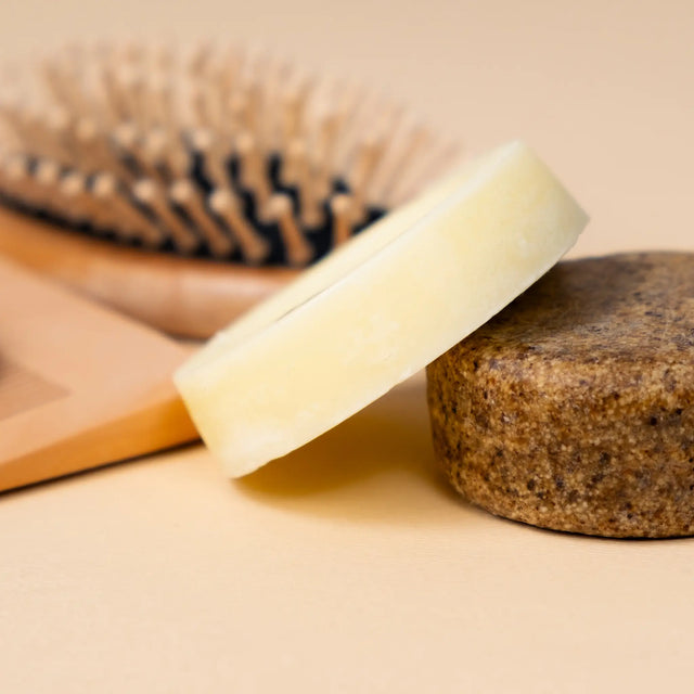 Twee ronde shampoobars op elkaar gestapeld met een houten haarborstel op de achtergrond – natuurlijke haarverzorging zonder plastic of siliconen
